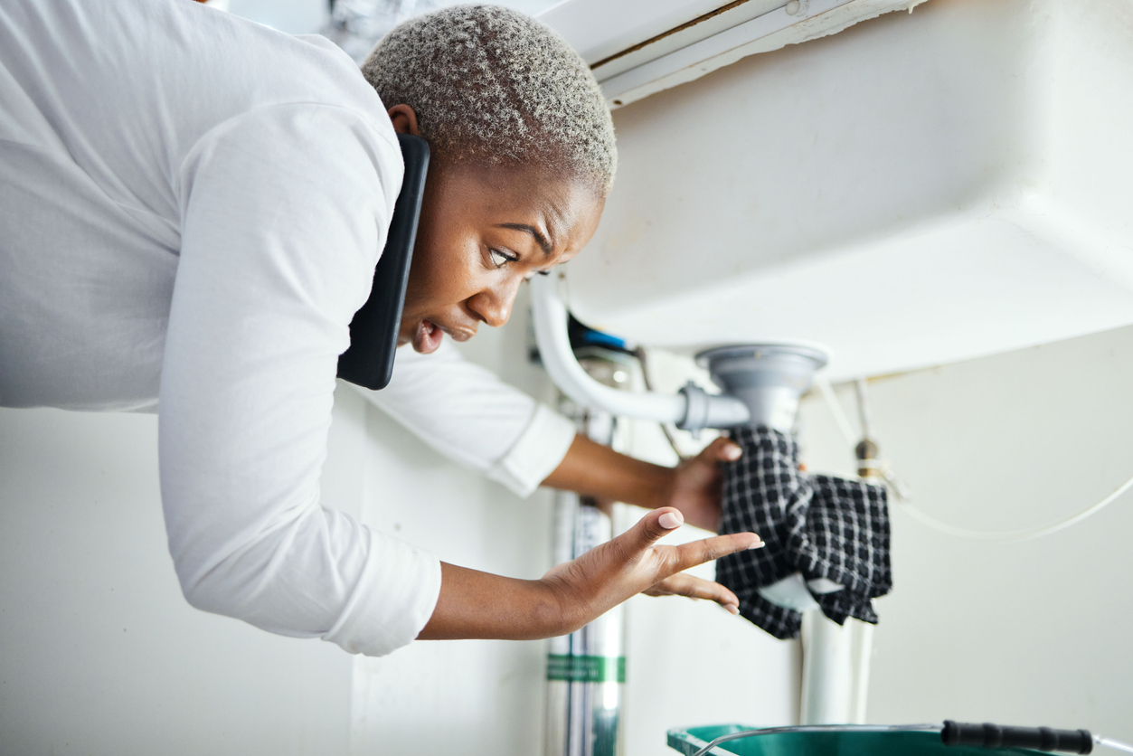 Homeowner checking inside a kitchen sink pipes on the phone with a plumber.